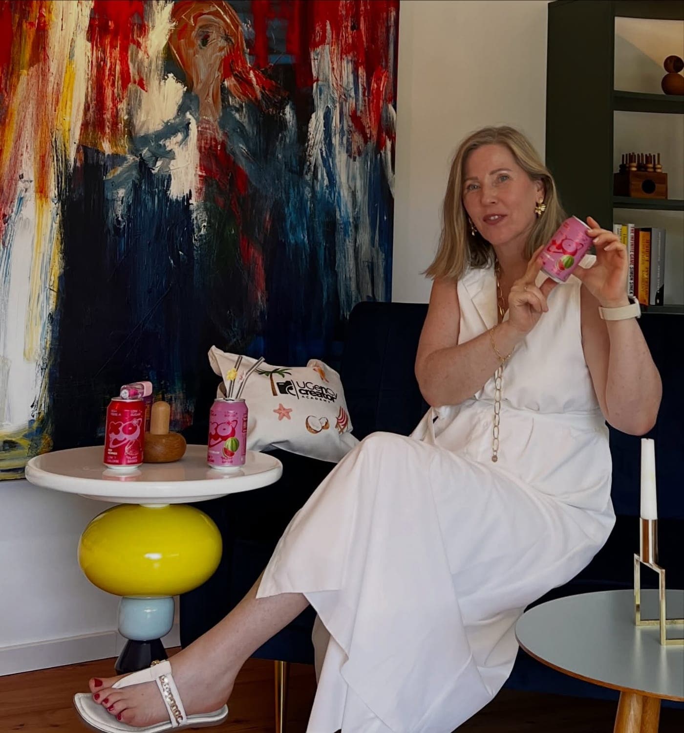 Dani sitzt in einem weißen Kleid auf einem blauen Sofa und zeigt eine Getränkedose