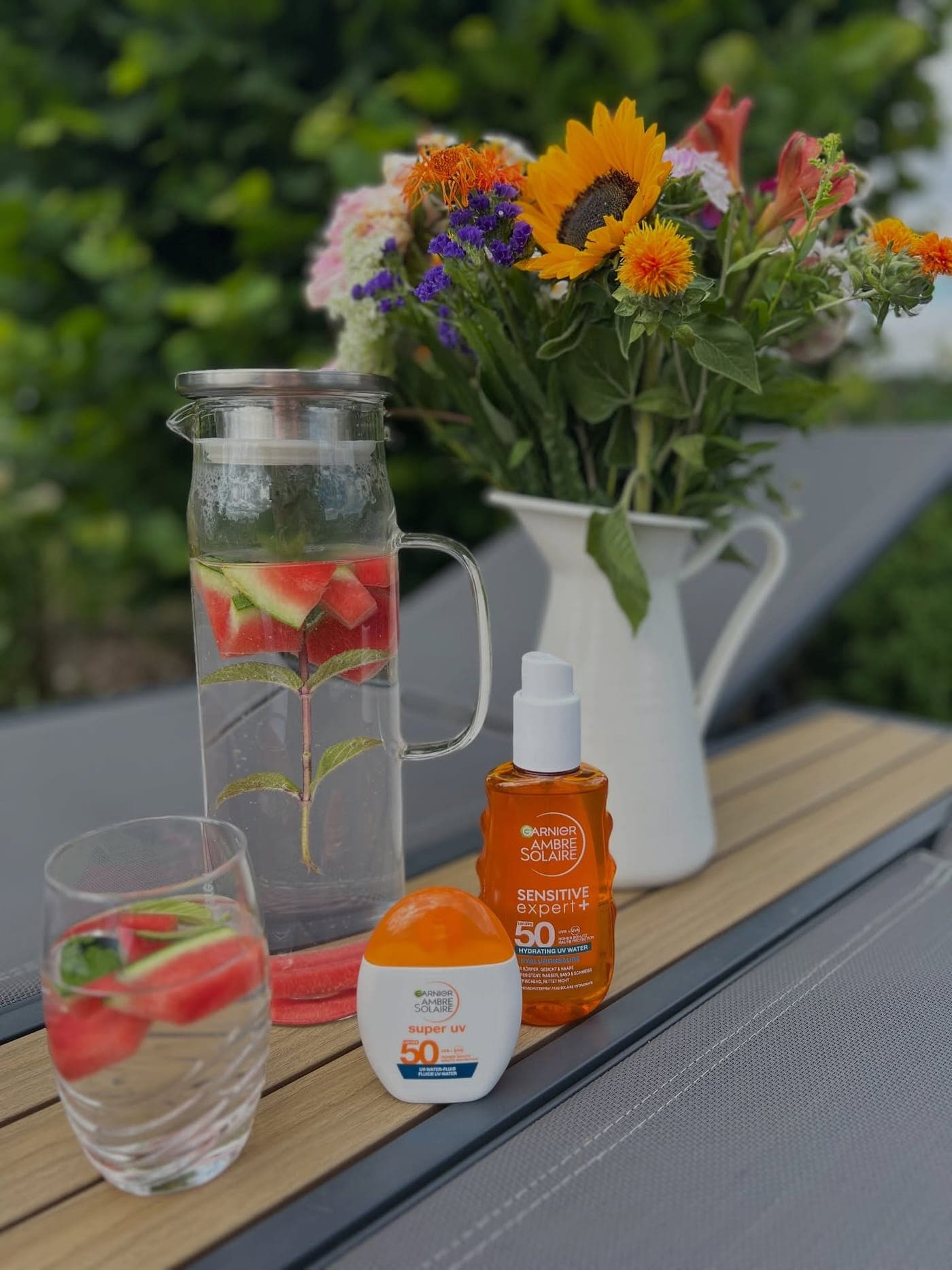 Karaffe mit Wassermelonenwasser, Sonnenpflege und ein Blumenstrauß auf einem Gartentisch